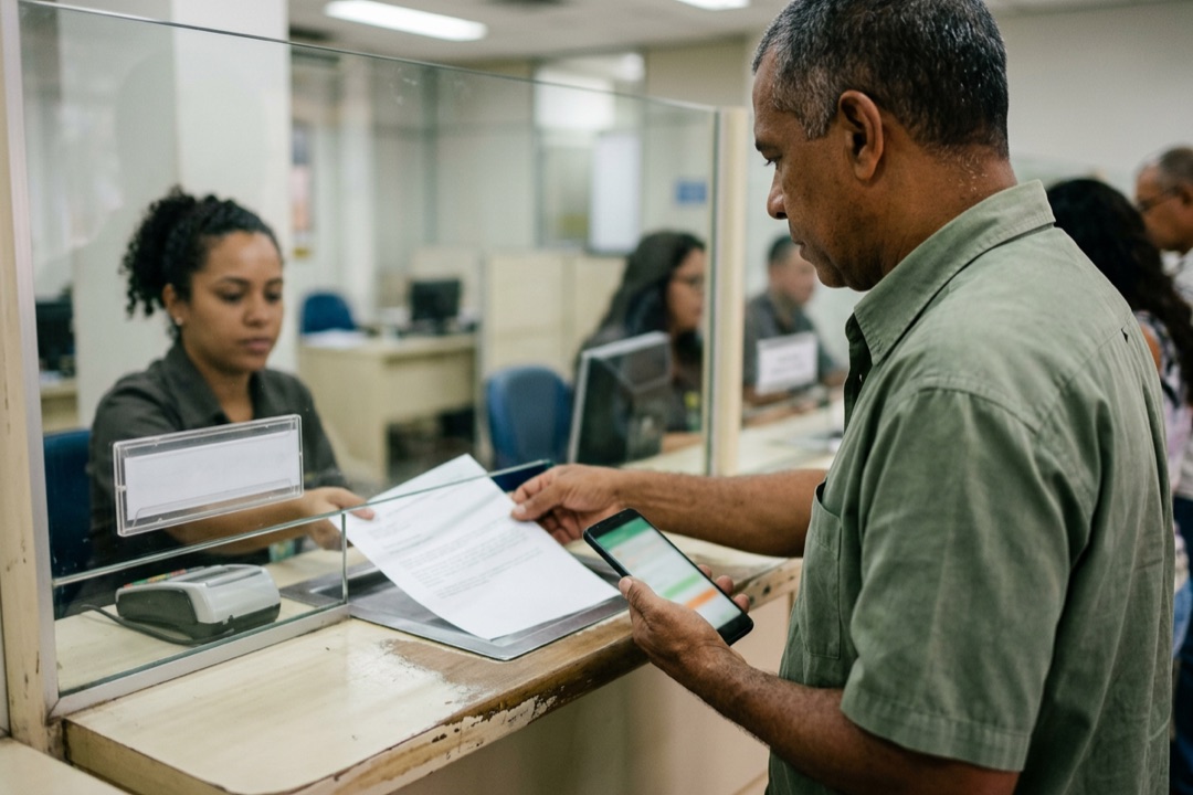 Homem entrega um comprovante impresso ao atendente no guichê de uma agência bancária