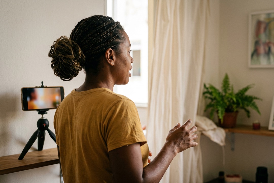 Mulher de uns 35 anos gravando um vídeo com o celular num tripé pequeno na sala de casa.