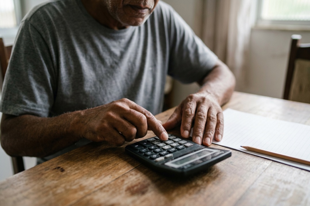 Mãos de um senhor usando uma calculadora simples ao lado de uma folha de papel e um lápis.