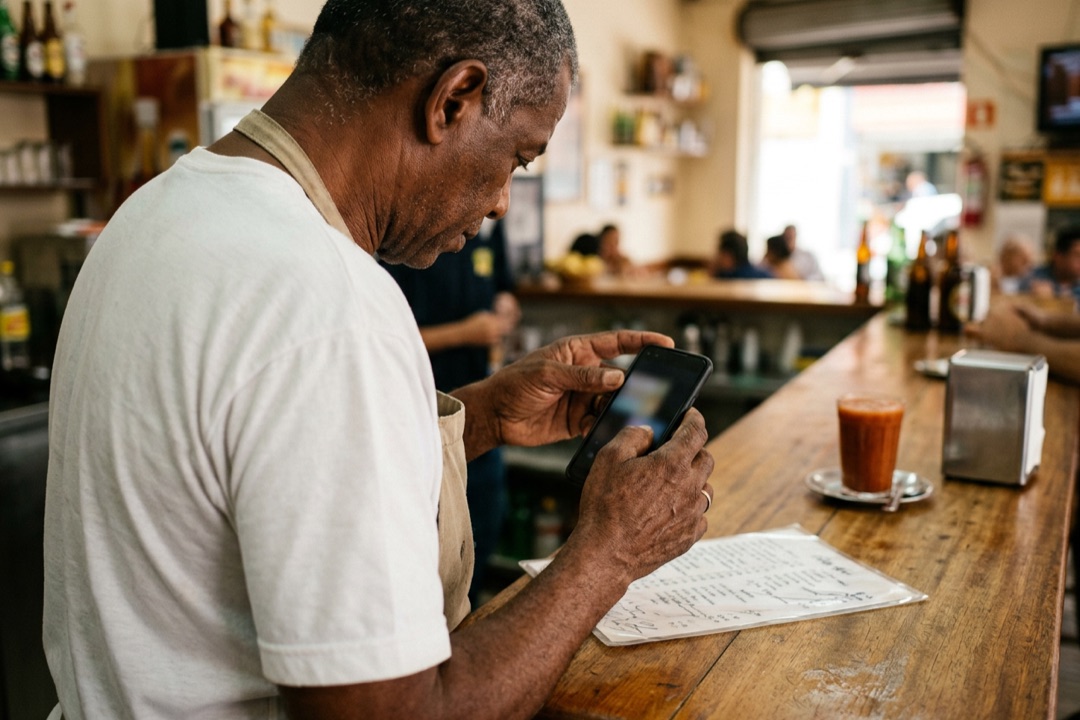 Dono de lanchonete fotografa com o celular o cardápio de papel sobre o balcão