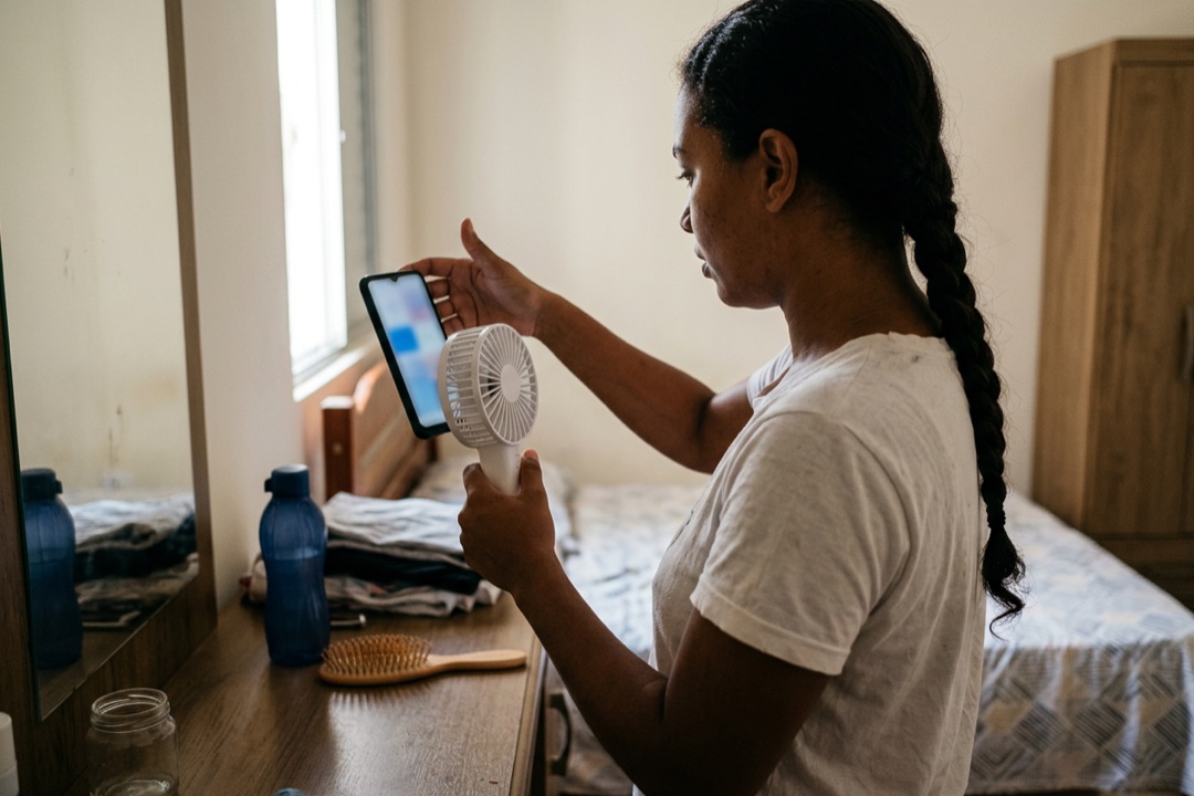 Mulher mostra um ventilador portátil branco de perto para a câmera do celular, no quarto, para gravar a demonstração.